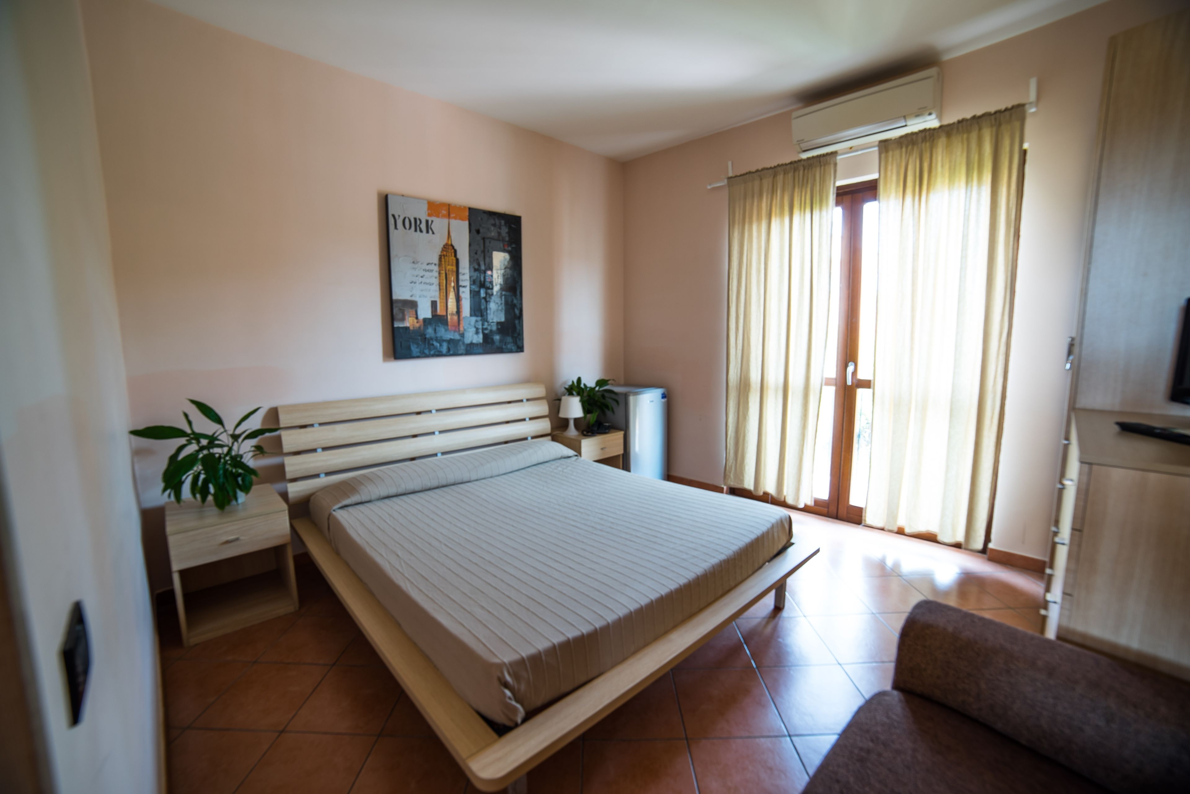 A bedroom with a bed, a nightstand, a lamp, a plant, a window with curtains, and a television.