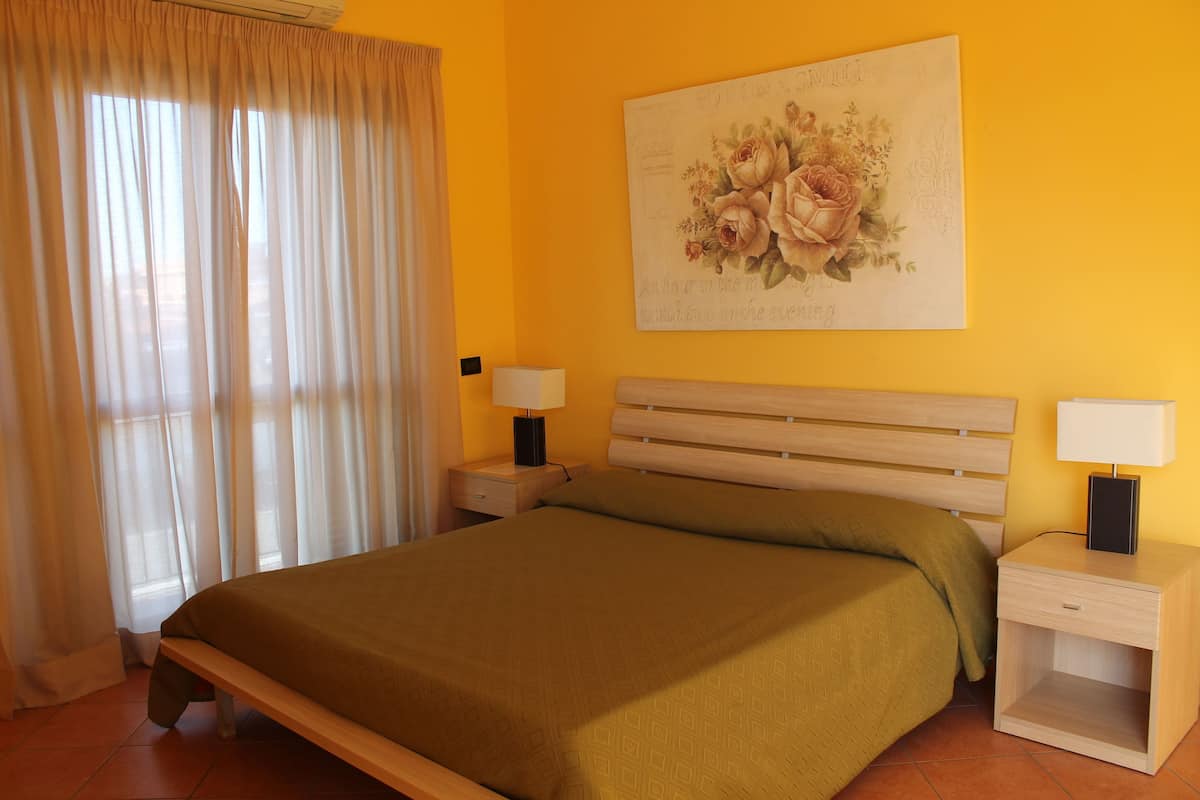 Ein Schlafzimmer mit einem Bett, zwei Nachttischen mit Lampen, einem Fenster mit Vorhängen und einem floralen Wandbild.