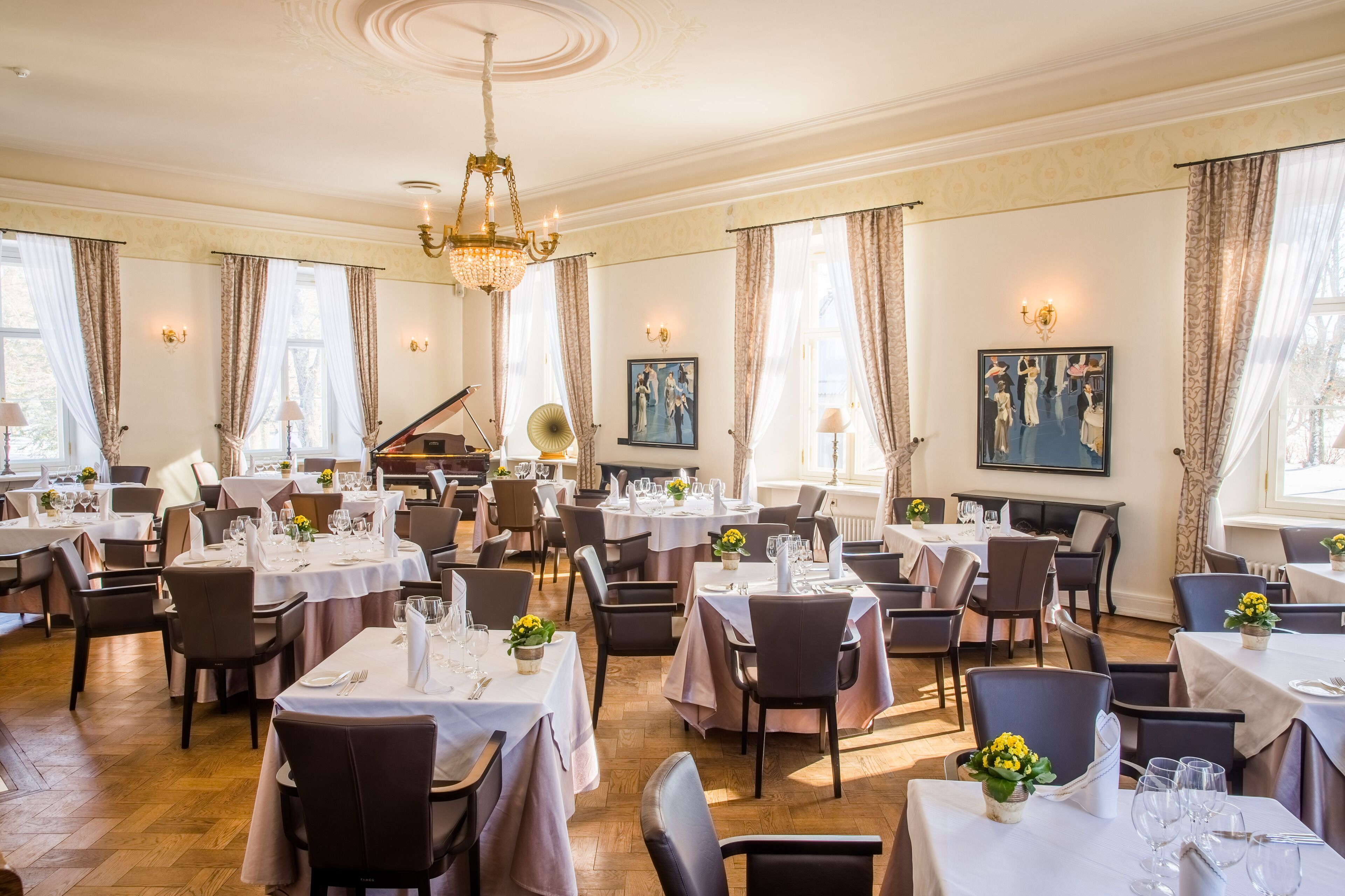 A formal dining room with multiple tables set for service, elegant chandeliers, and large windows with curtains.