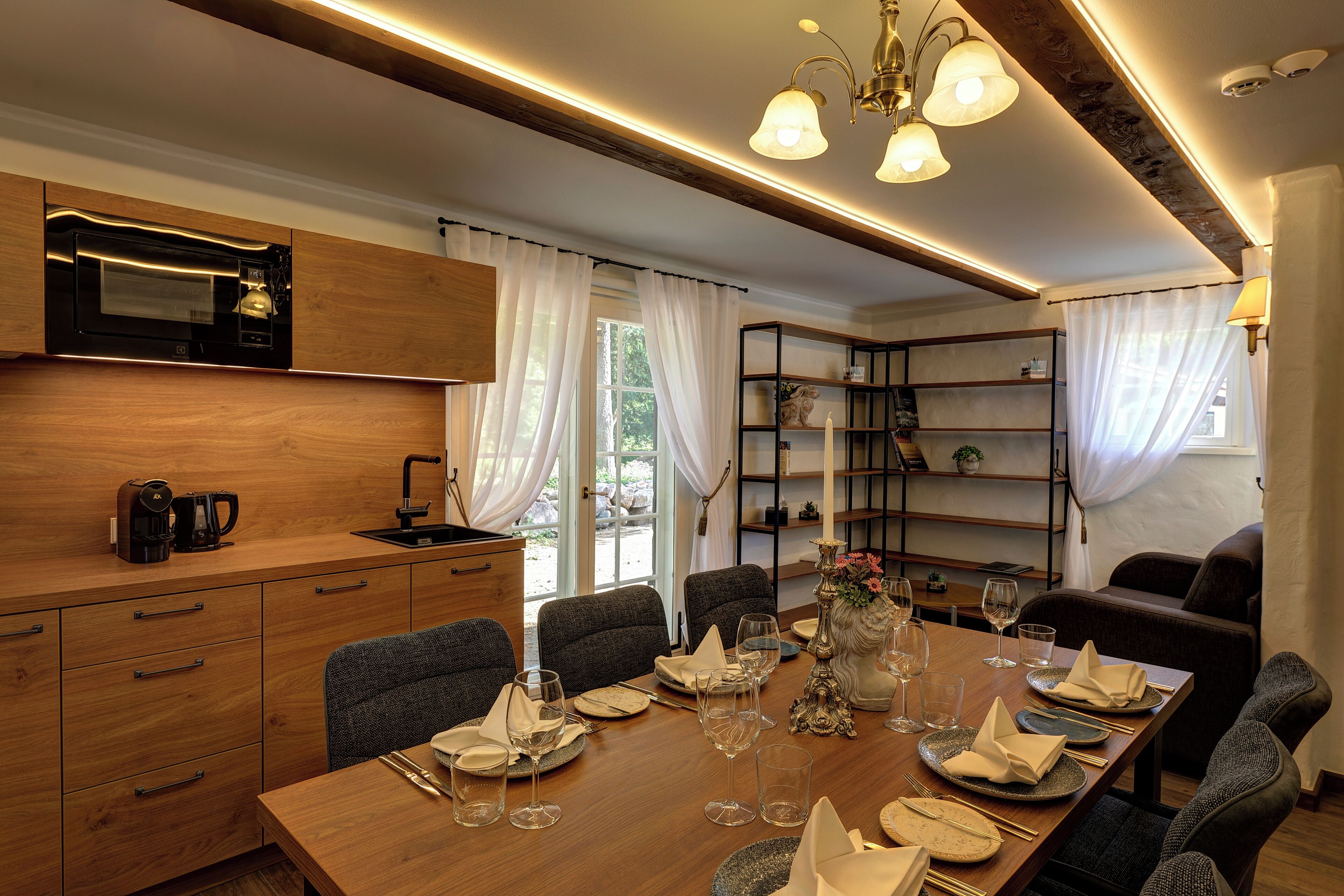 Un comedor con una mesa de madera dispuesta para cuatro personas, un microondas sobre la encimera y una estantería con objetos decorativos.