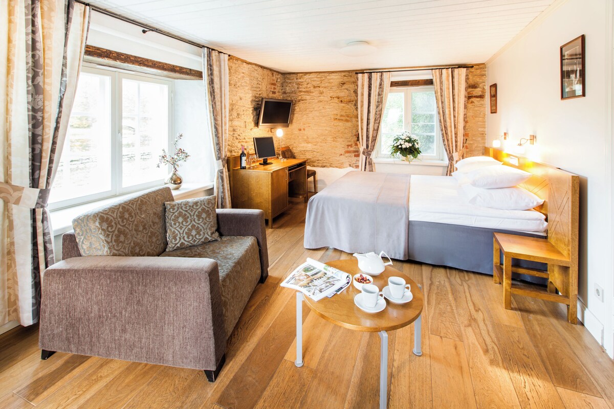 A hotel room with a sofa, a bed, a desk with a computer, a small table with cups and a newspaper, and a window with curtains.