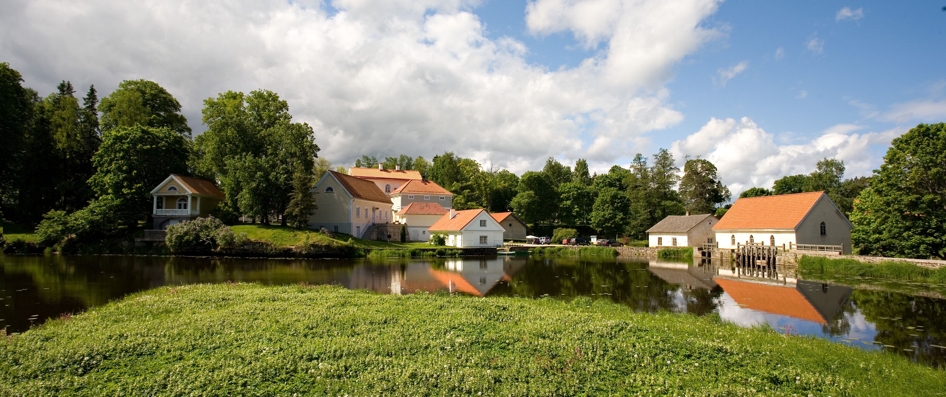 Una escena apacible junto al río con edificios tradicionales, una masa de agua tranquila y abundante vegetación.