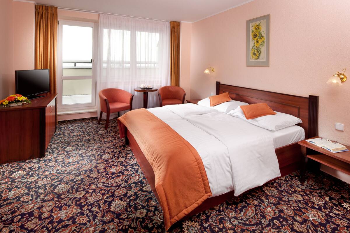Une chambre d’hôtel avec un grand lit, deux chaises, un bureau avec une télévision, une petite table et une fenêtre avec des rideaux.