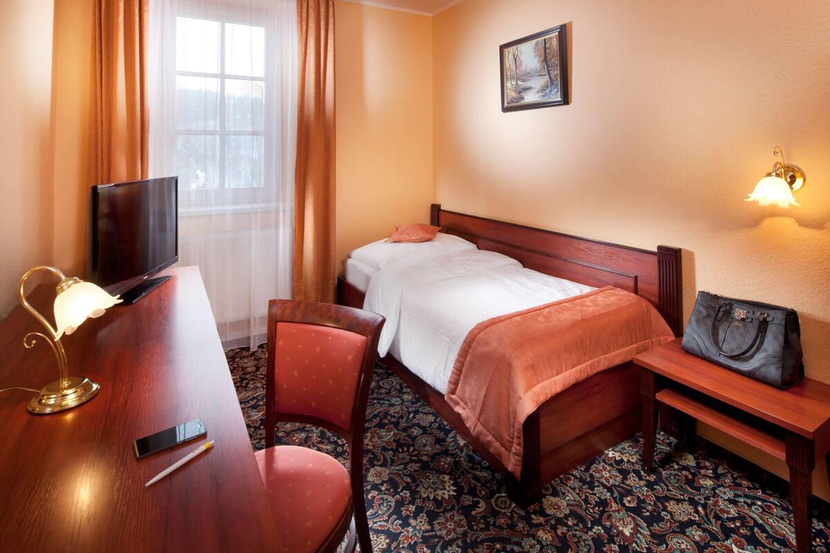 Une chambre d’hôtel avec un lit, un bureau avec une lampe, une chaise, une table de chevet avec un sac à main, et un tableau accroché au mur.