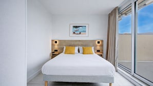 Habitación de hotel moderna con una cama grande, dos mesitas de noche con lámparas, un cuadro en la pared y un ventanal con cortinas.