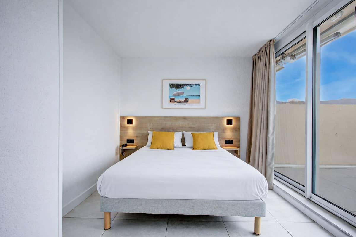 A modern hotel room with a large bed, two bedside tables with lamps, a painting on the wall, and a large window with curtains.