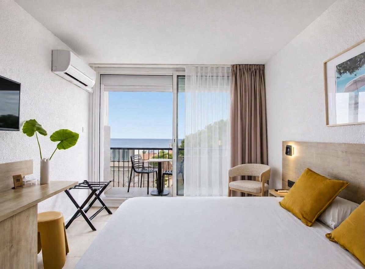 A hotel room with a bed, a desk, a chair, a plant, and a balcony with a view.