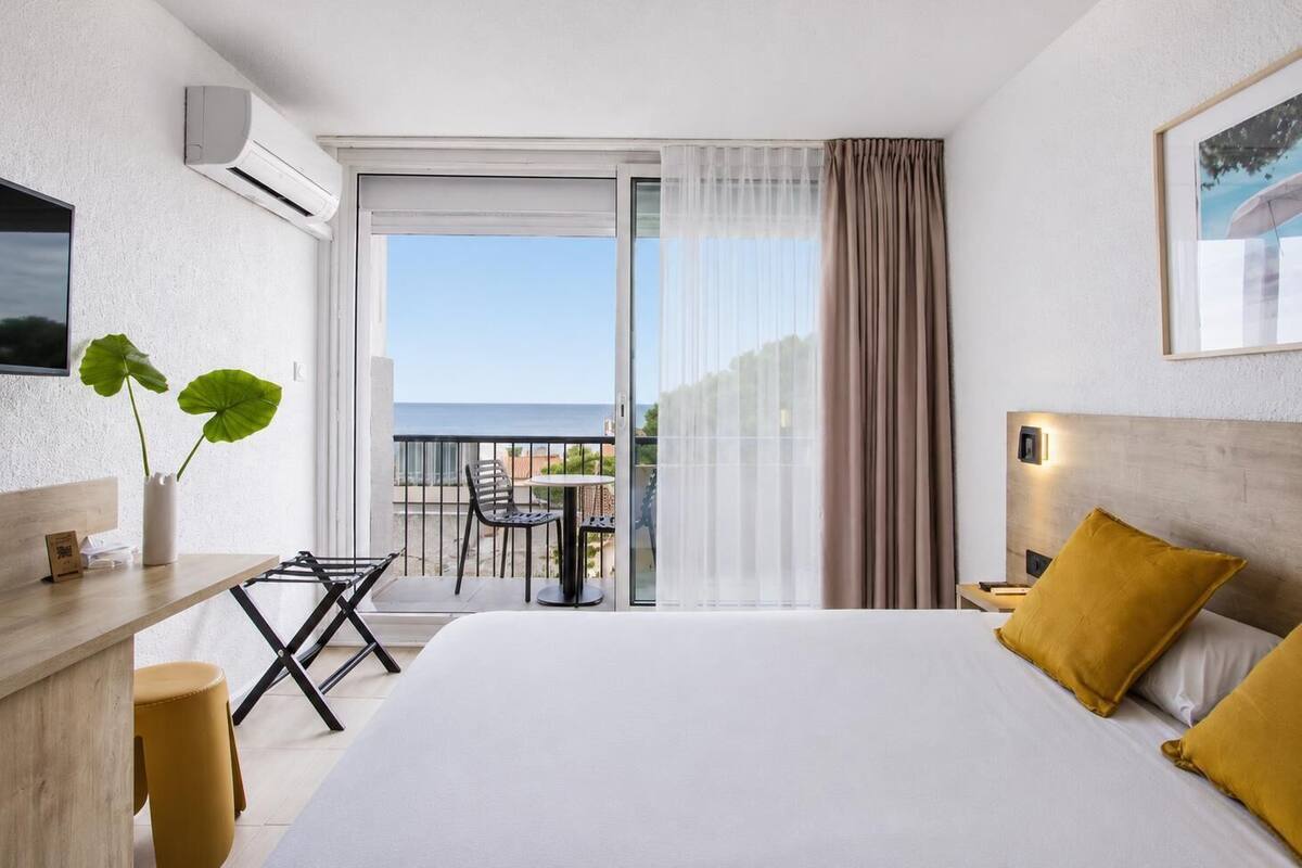 A hotel room with a bed, a desk with a chair, a small table with chairs, a potted plant, and a balcony with a view.