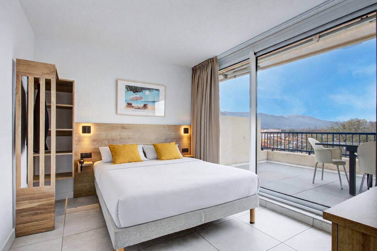 A modern hotel room with a large bed, a wooden wardrobe, a bedside table with a lamp, a framed picture on the wall, and a balcony with a view of the mountains.