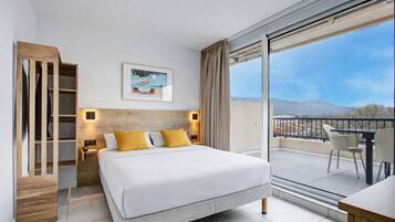 Habitación de hotel moderna con una cama grande, un armario de madera, una mesita de noche con una lámpara, un cuadro en la pared y un balcón con vistas a las montañas.