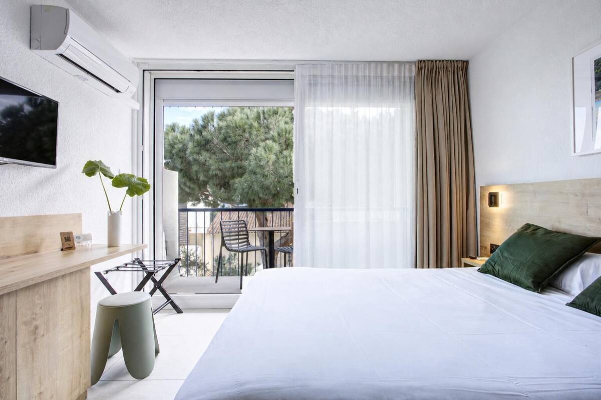 A hotel room with a bed, a TV, a desk, a chair, a plant, and a balcony with a view of trees.