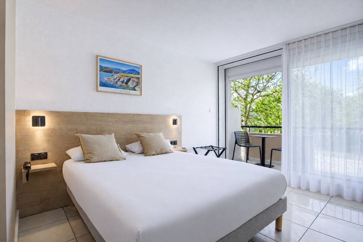 A hotel room with a large bed, a small table with chairs, a balcony with a view, and a painting on the wall.