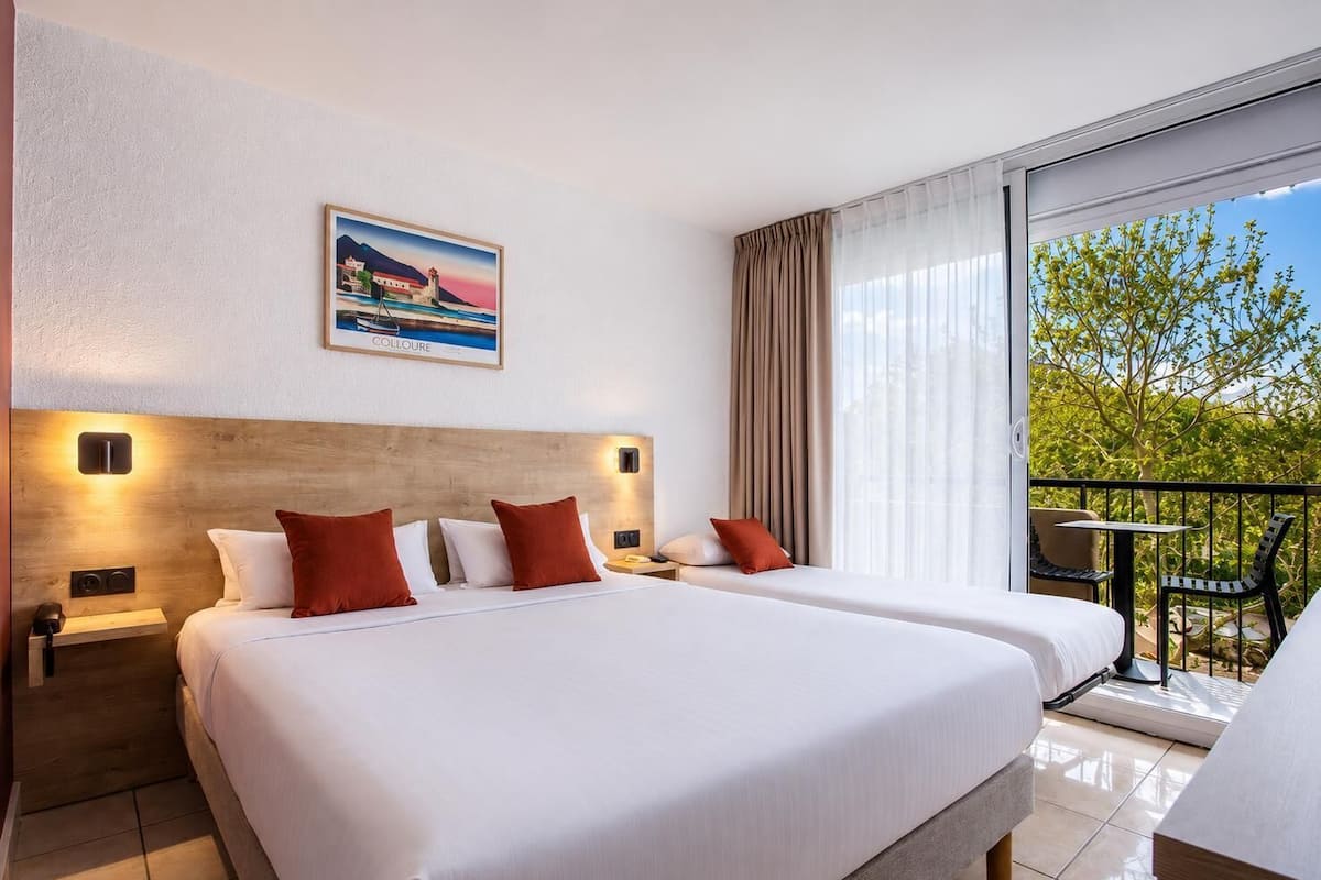 A hotel room with two beds, a painting, a balcony with a table and chairs, and a view of greenery.