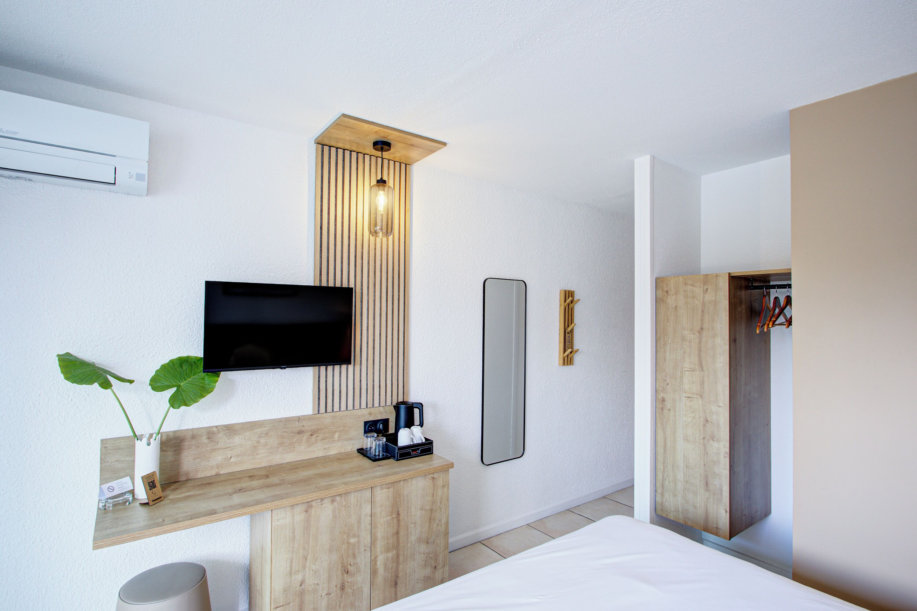 Una habitación de hotel moderna con un escritorio de madera, un televisor de pantalla plana, una mesita de noche, un espejo y un armario empotrado.