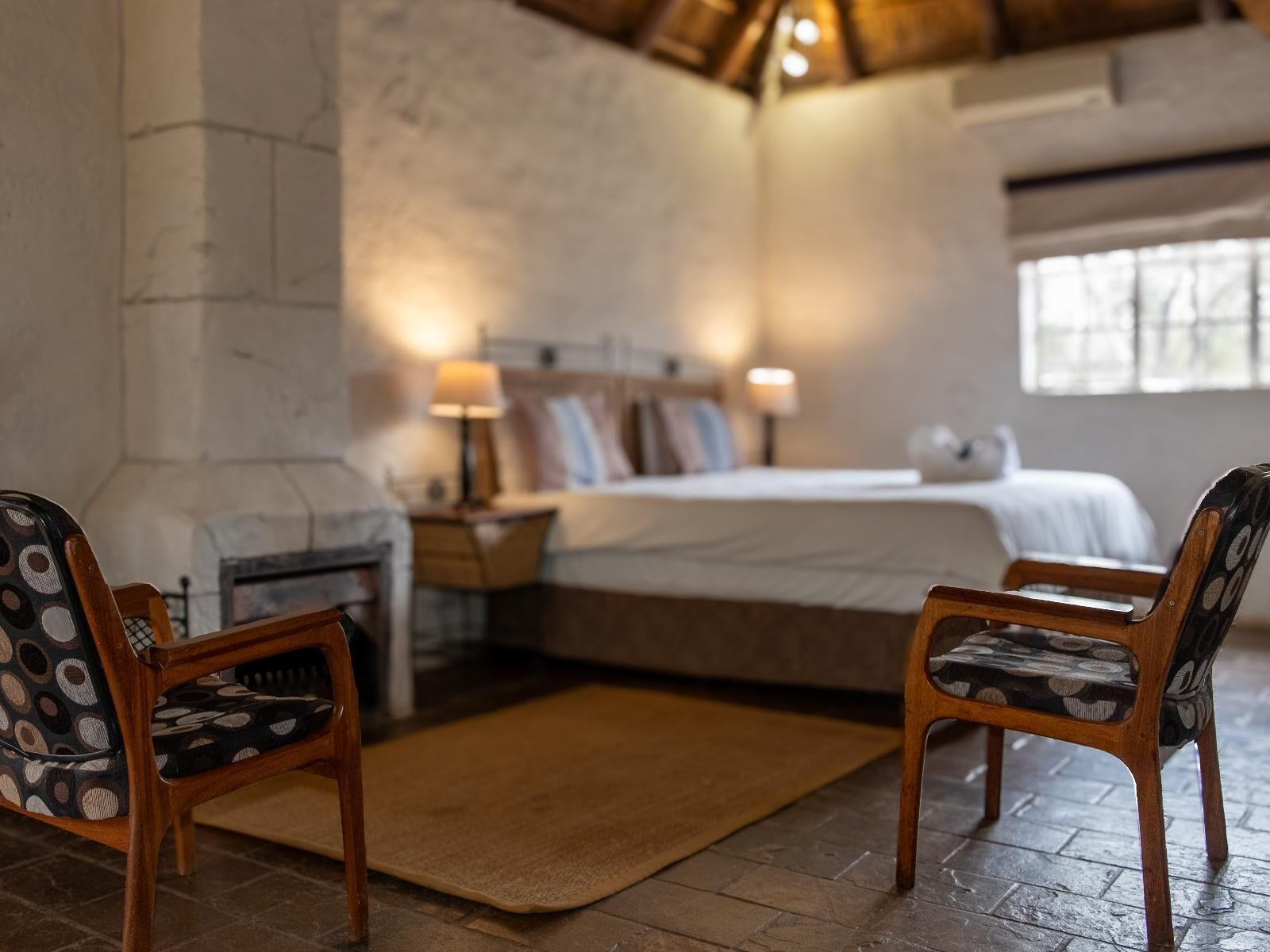Un dormitorio acogedor con una cama grande, dos sillas de madera con cojines estampados, una mesita de noche con lámpara y una chimenea de piedra.