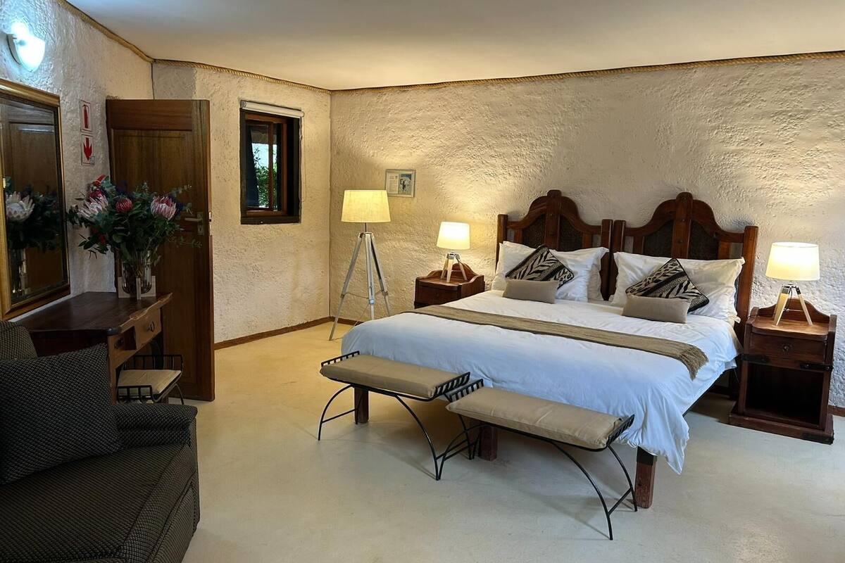 A bedroom with a bed, bedside tables, a bench, a chair, and a desk with a lamp and flowers.