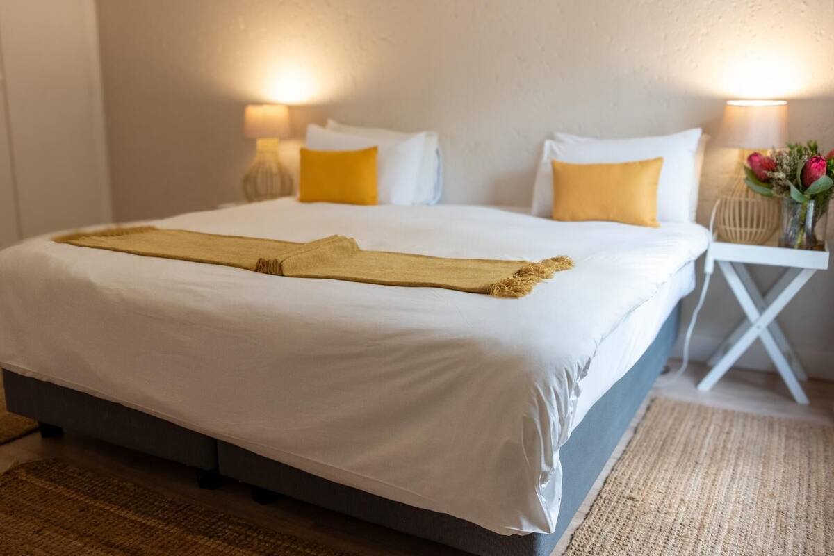 A neatly made bed with white linens and a beige throw, flanked by two white pillows and two yellow pillows. There are two bedside lamps with white shades, and a small table with a vase of flowers to the right.