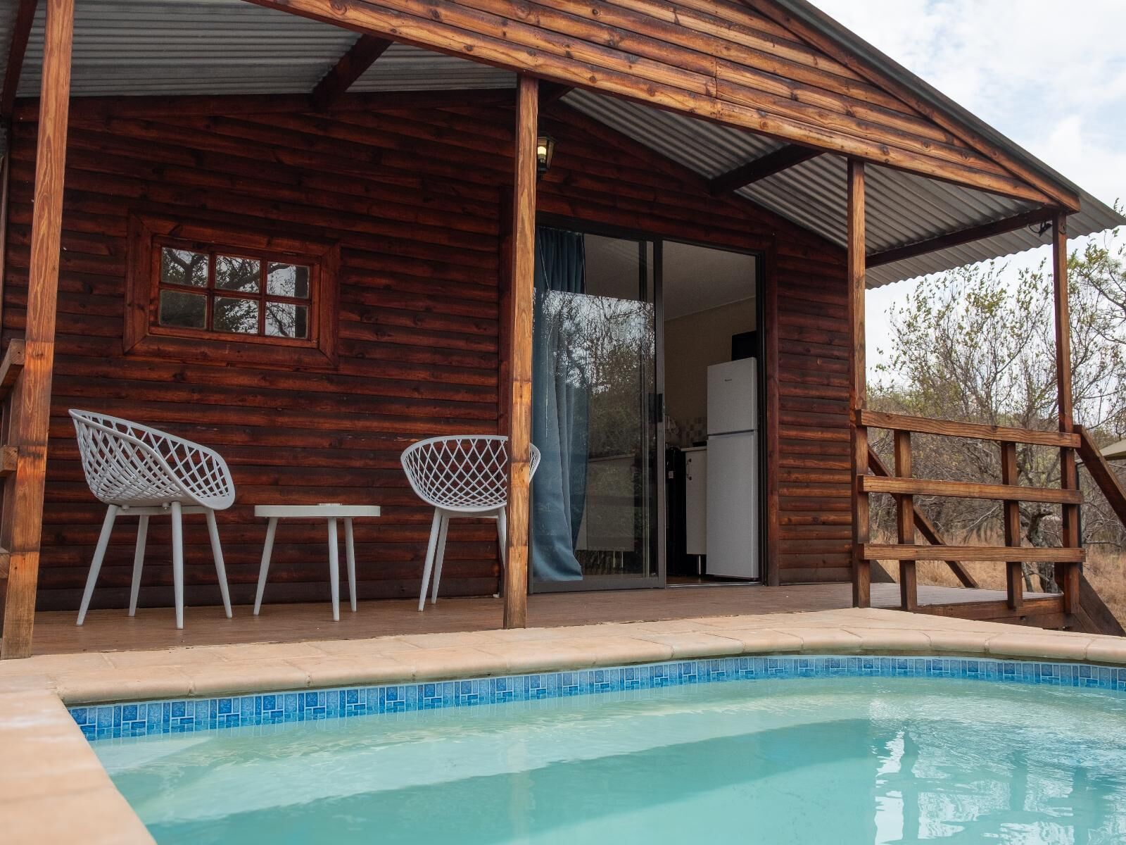 Una cabaña de madera con piscina, dos sillas blancas y un refrigerador en su interior.