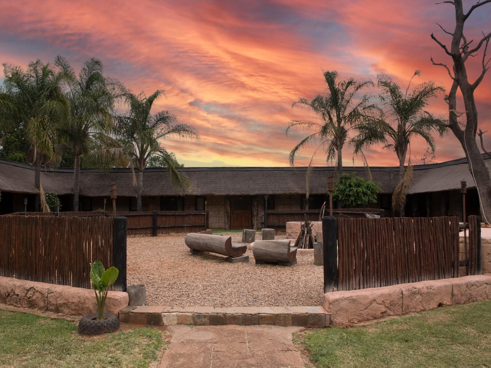 A courtyard with thatched-roof huts, surrounded by wooden fences and palm trees, under a sunset sky.