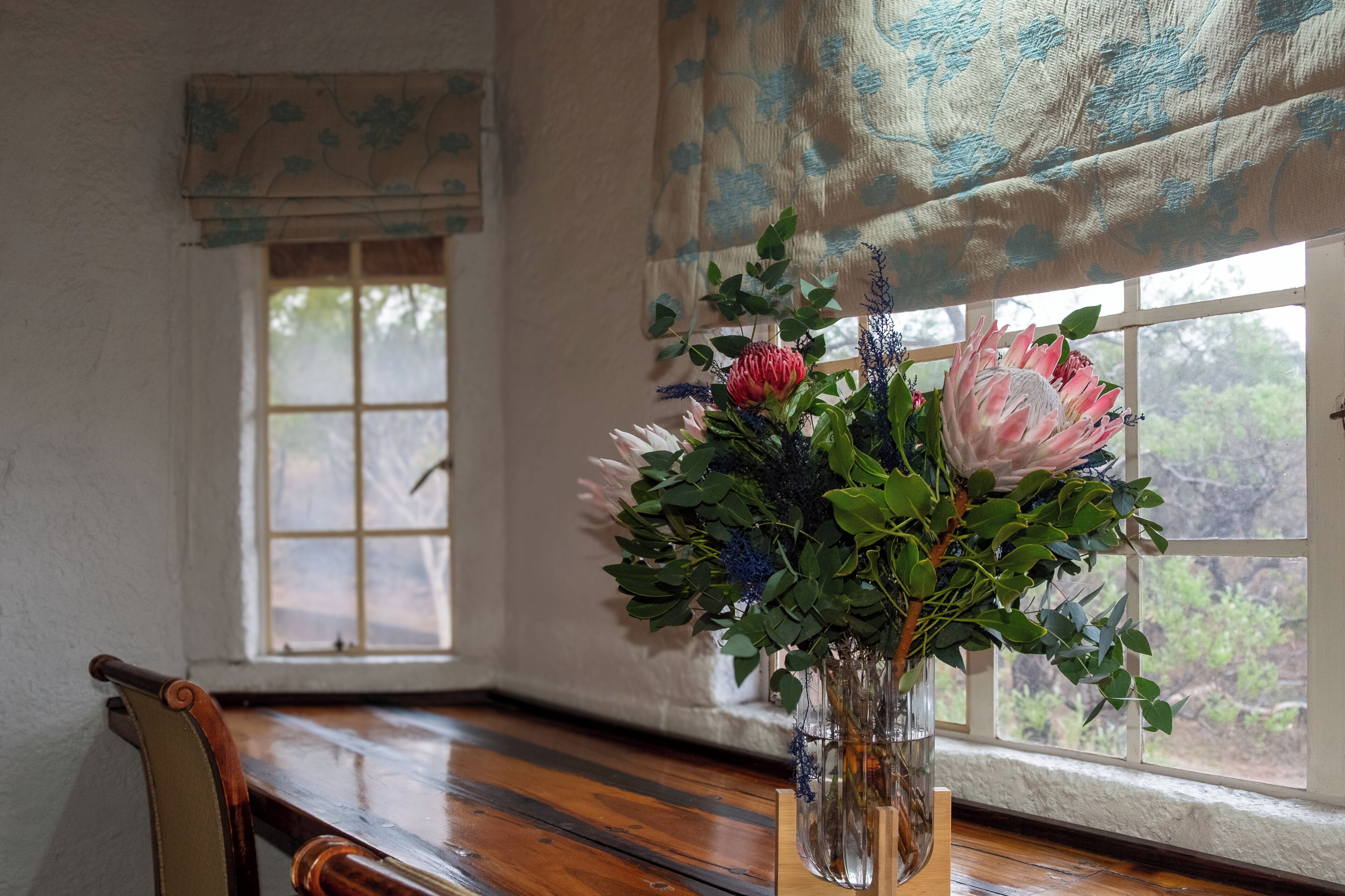 Una mesa de madera con un jarrón de flores frente a una ventana que da a árboles.
