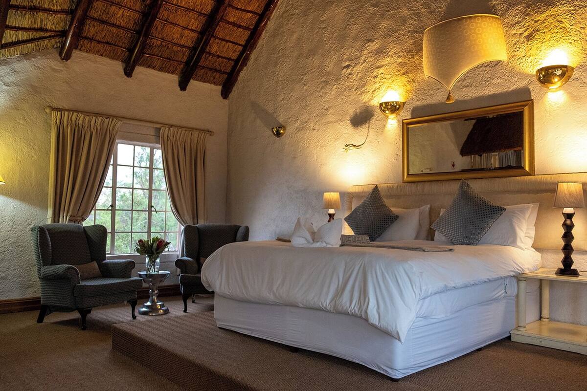 A bedroom with a large bed, two chairs, a small table with a vase, and a window with curtains.