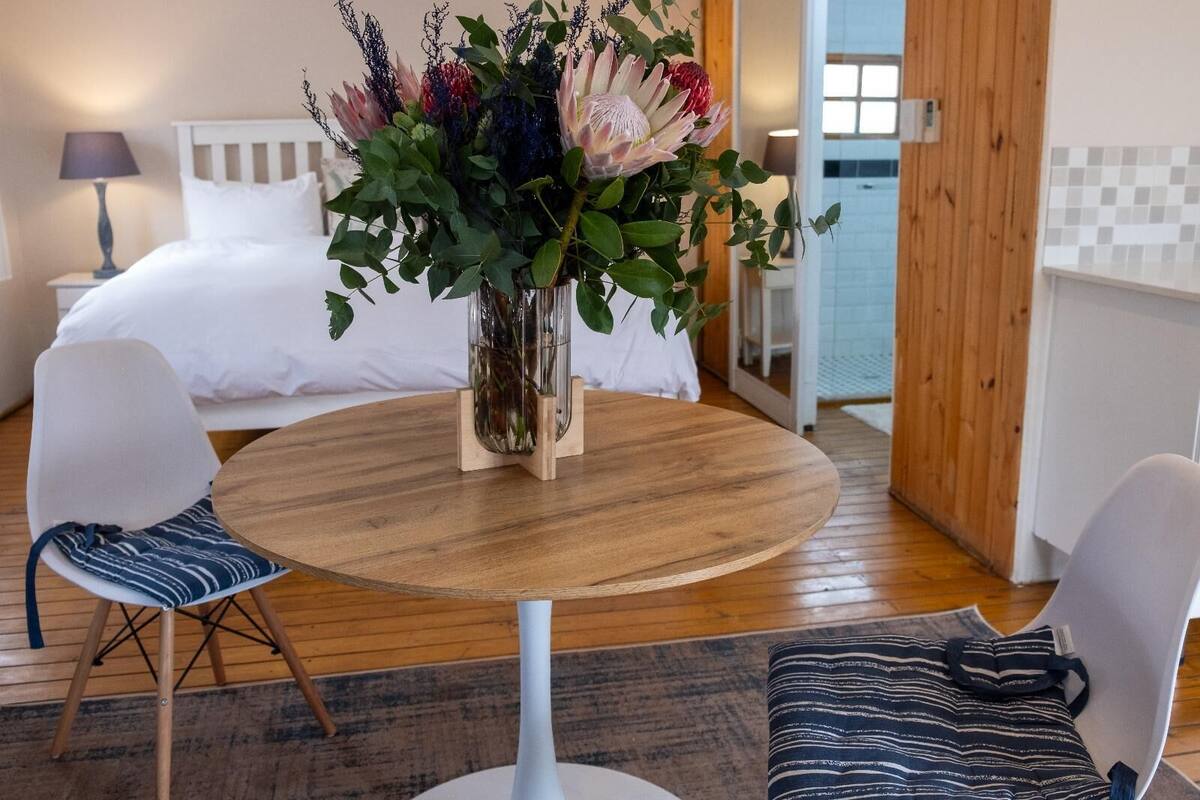 A hotel room with a round wooden table, two white chairs, a bed with white bedding, and a large floral arrangement on the table.