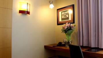 Habitación de hotel con escritorio, teléfono, un cuadro enmarcado, un jarrón con flores y una lámpara.