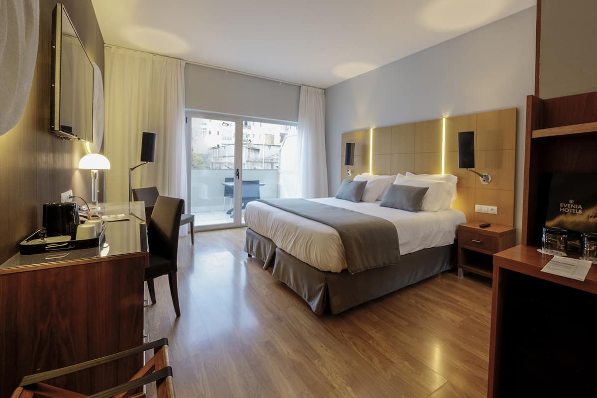A hotel room with a large bed, a desk with a lamp, a chair, and a view of the outside through a sliding door.