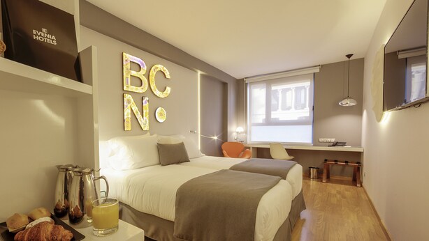 A hotel room with a bed, bedside table with a coffee pot and croissant, a desk with a chair, and a window with a view. - Evenia Rossello (Barcelona)