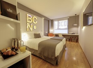 A hotel room with a bed, bedside table with a coffee pot and croissant, a desk with a chair, and a window with a view. - Evenia Rossello (Barcelona)
