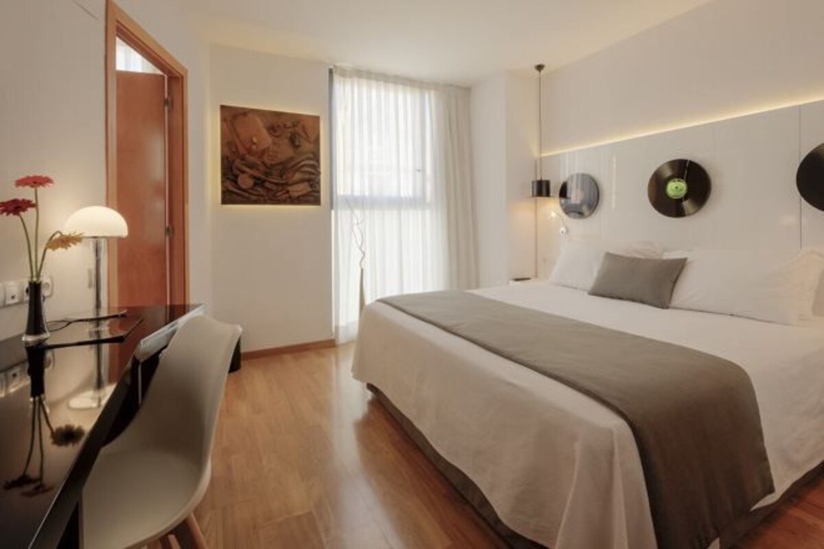 Quarto de hotel com cama, mesa de cabeceira, luminária, poltrona e uma obra de arte na parede.