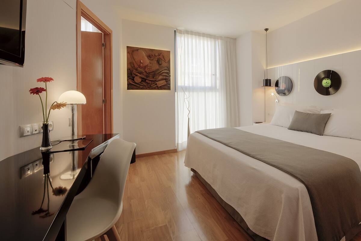 A hotel room with a bed, bedside tables, a desk, a chair, a lamp, a painting, and a window with curtains.