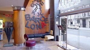 A modern lobby with a wooden wall displaying the name 'BARCELONA' in large letters, a seating area with a sofa and chairs, and a glass entrance to the street. - Evenia Rossello (Barcelona)