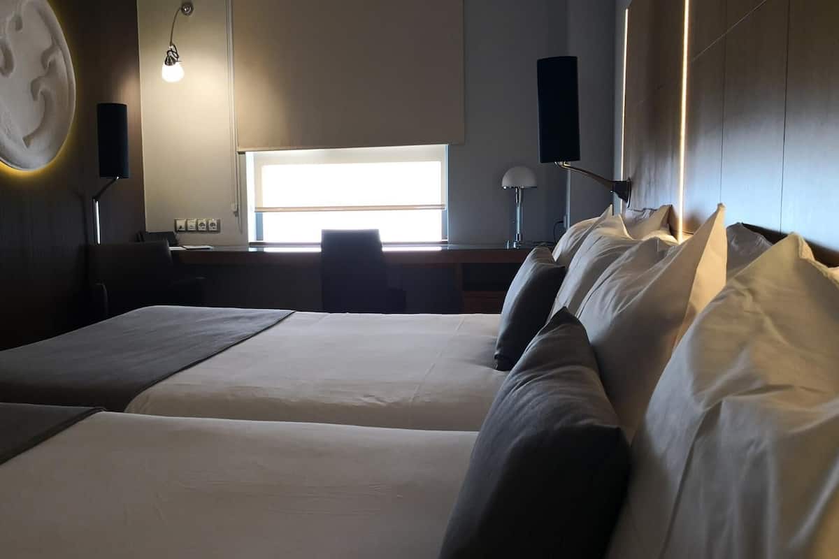 A hotel room with two beds, a desk, a chair, a window, and wall-mounted lamps.