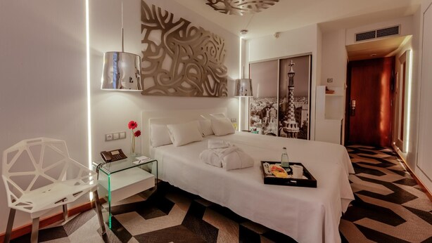 A modern hotel room with a large bed, a glass side table, a decorative wall panel, and a view of the city through the window. - Evenia Rossello (Barcelona)