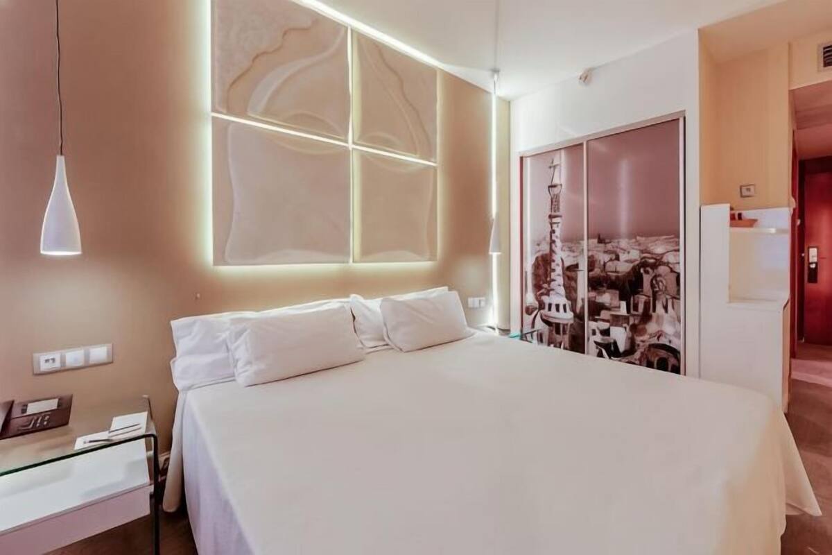 A modern hotel room with a large bed, a bedside table, a wardrobe, and a wall-mounted light fixture.