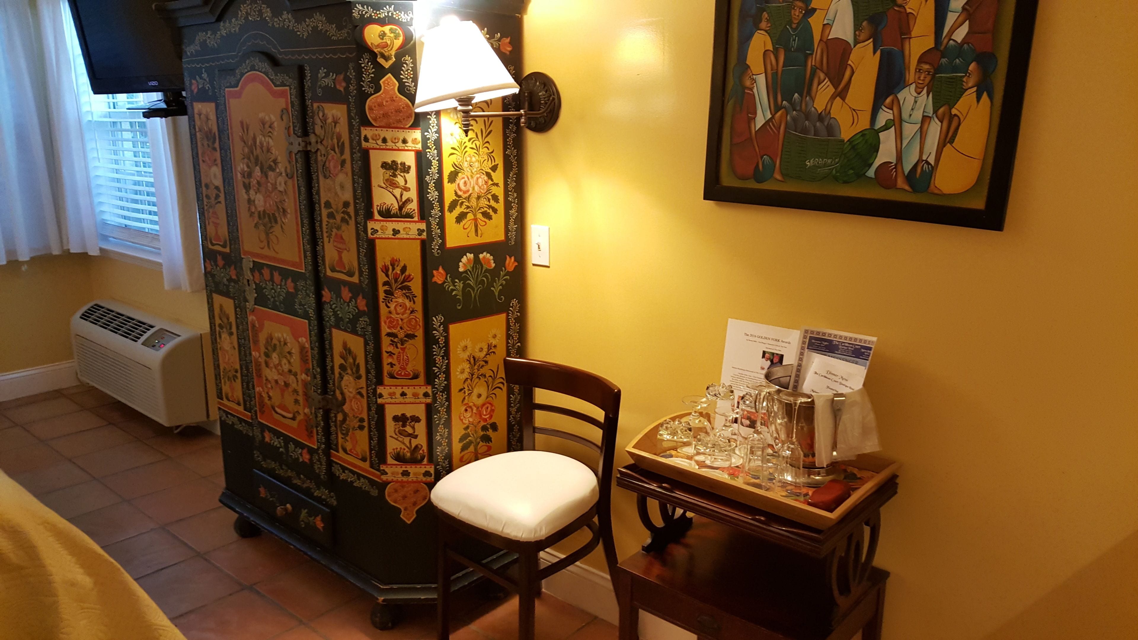 Ein Zimmer mit einem traditionellen Schrank, einem Sessel, einem Tisch mit Gläsern und einem gerahmten Gemälde an der Wand.
