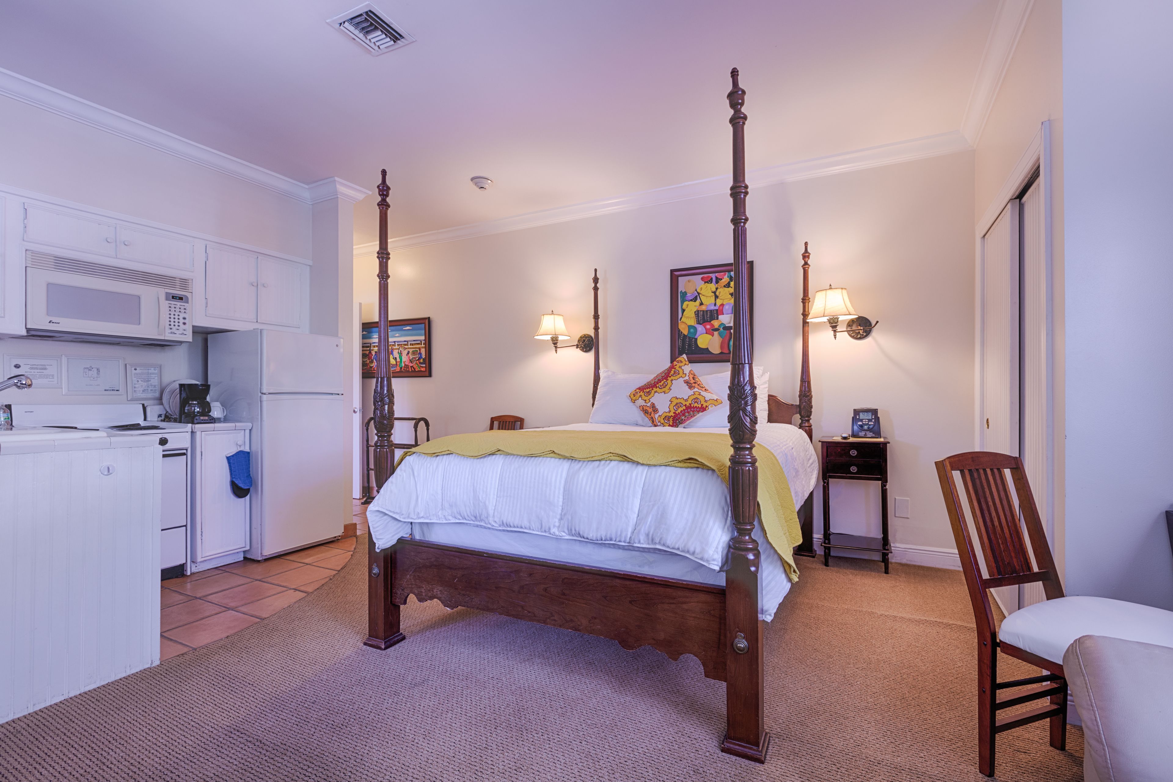 A four-poster bed with a wooden frame, a small bedside table with a lamp, a chair, and a kitchenette with a microwave and refrigerator.