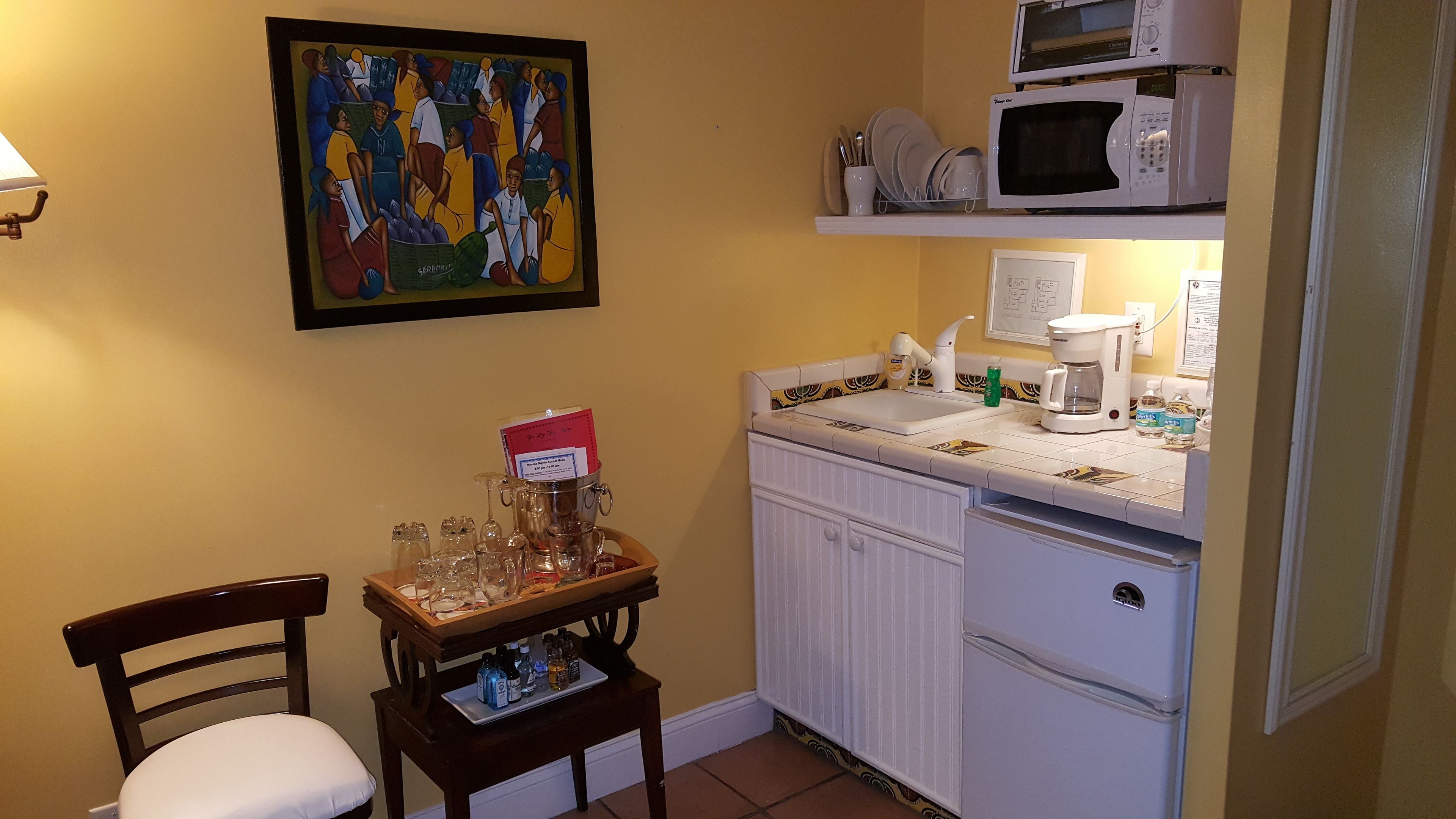 Una cocina pequeña con microondas, cafetera y una estantería con vajilla. Hay una silla y una mesa con vasos y botellas.