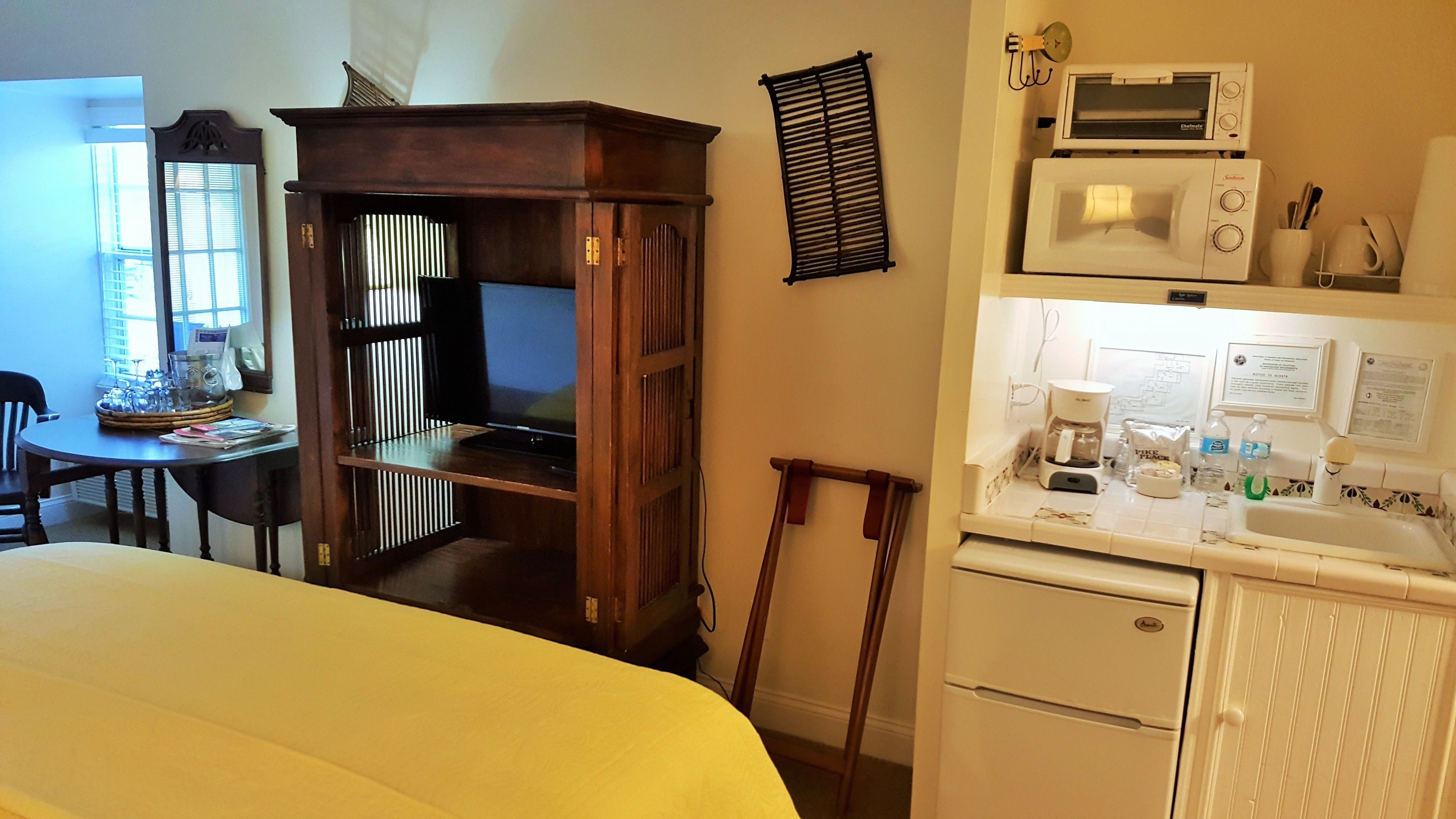 A compact hotel room with a bed, a small desk with a chair, a wooden wardrobe, a kitchenette with a microwave, coffee maker, and a sink.
