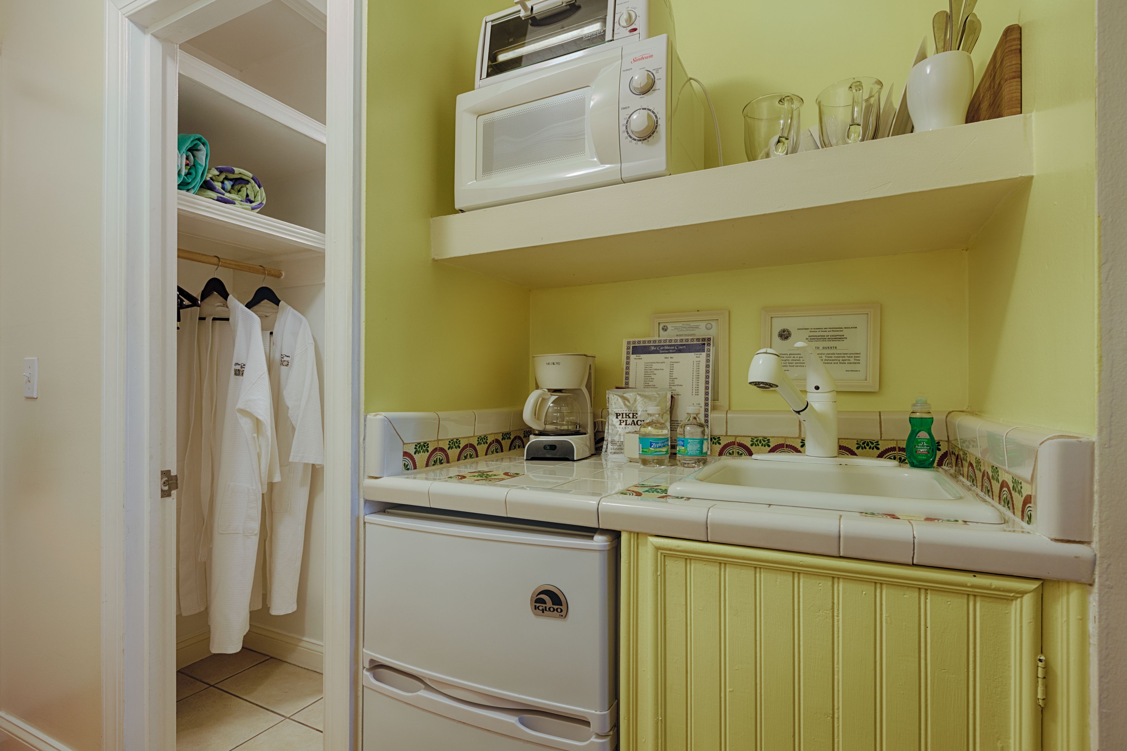 A compact kitchen with a microwave, coffee maker, and a sink. There is a small refrigerator and a cabinet with a stove.