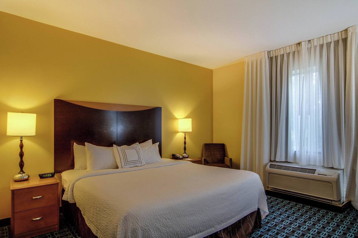 A hotel room with a large bed, two bedside lamps, a chair, a bedside table with a phone, and a window with curtains.