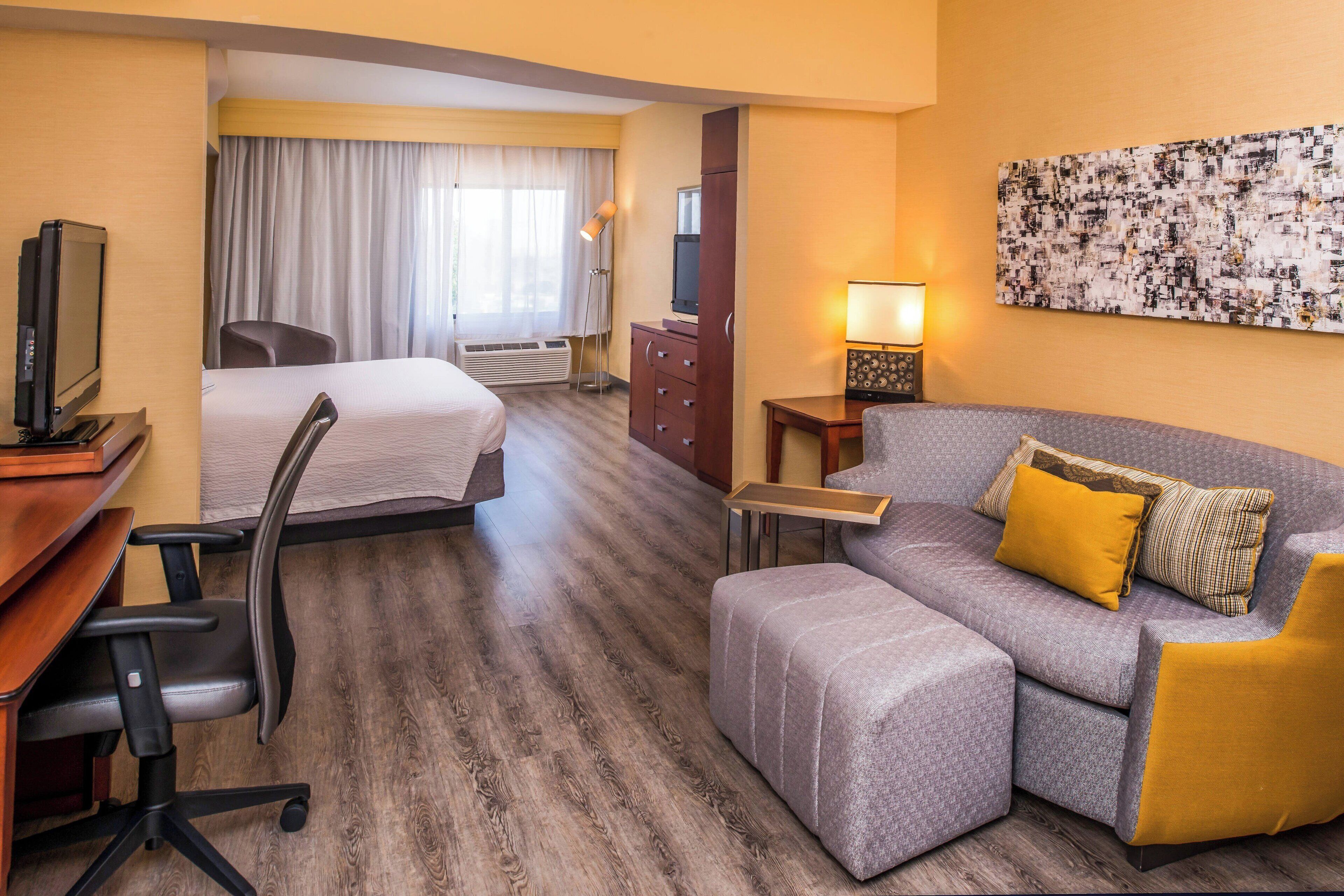 A hotel room with a bed, a sofa, a desk with a computer, a TV, a lamp, and a painting on the wall.