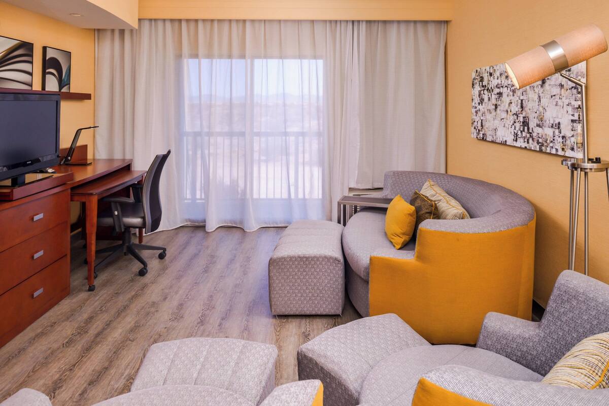 A hotel room with a wooden desk, a television, a sofa with cushions, a large window with curtains, and a floor lamp.