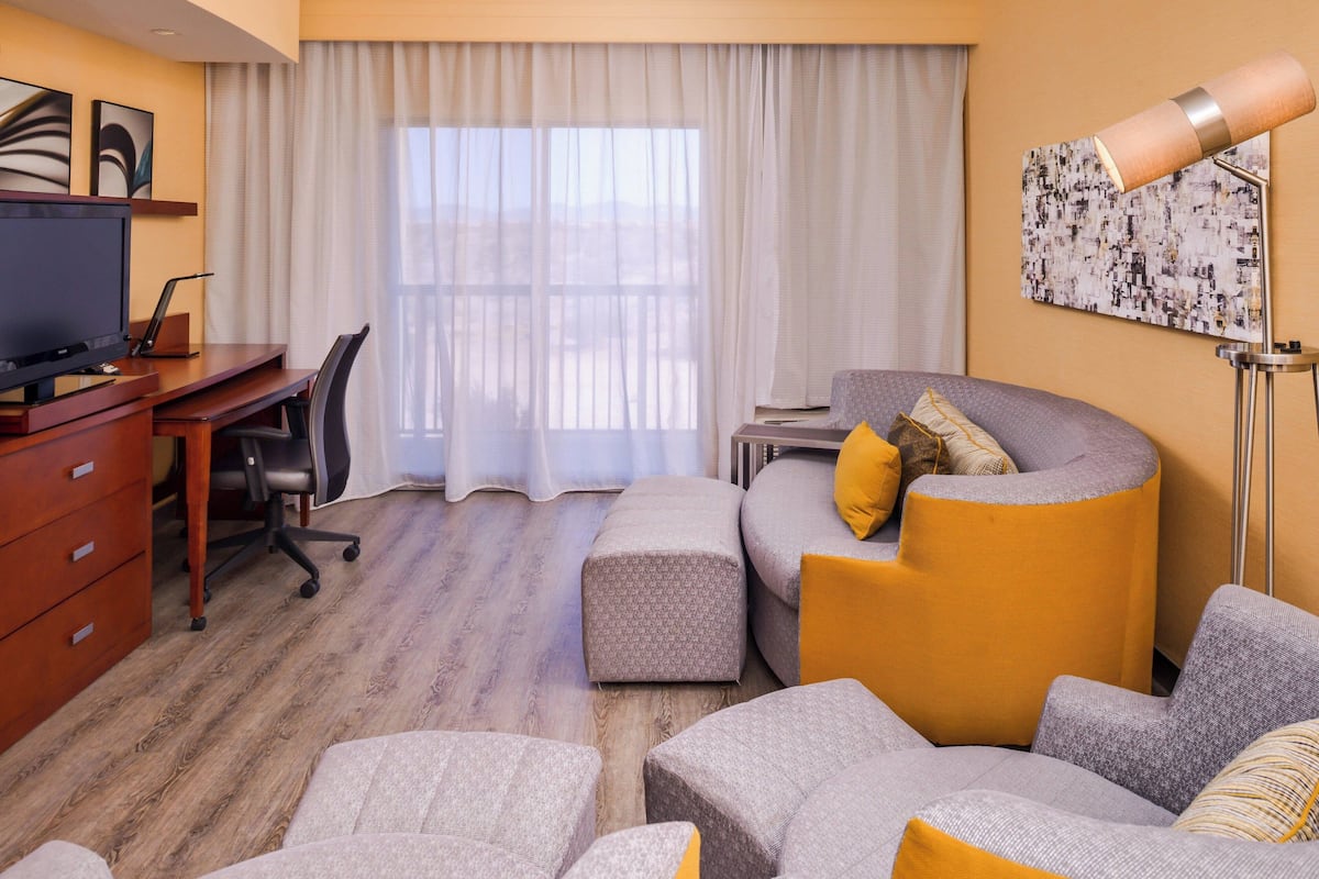 Une chambre d’hôtel avec un bureau en bois, une télévision, un canapé avec des coussins, une grande fenêtre avec des rideaux et une lampe sur pied.