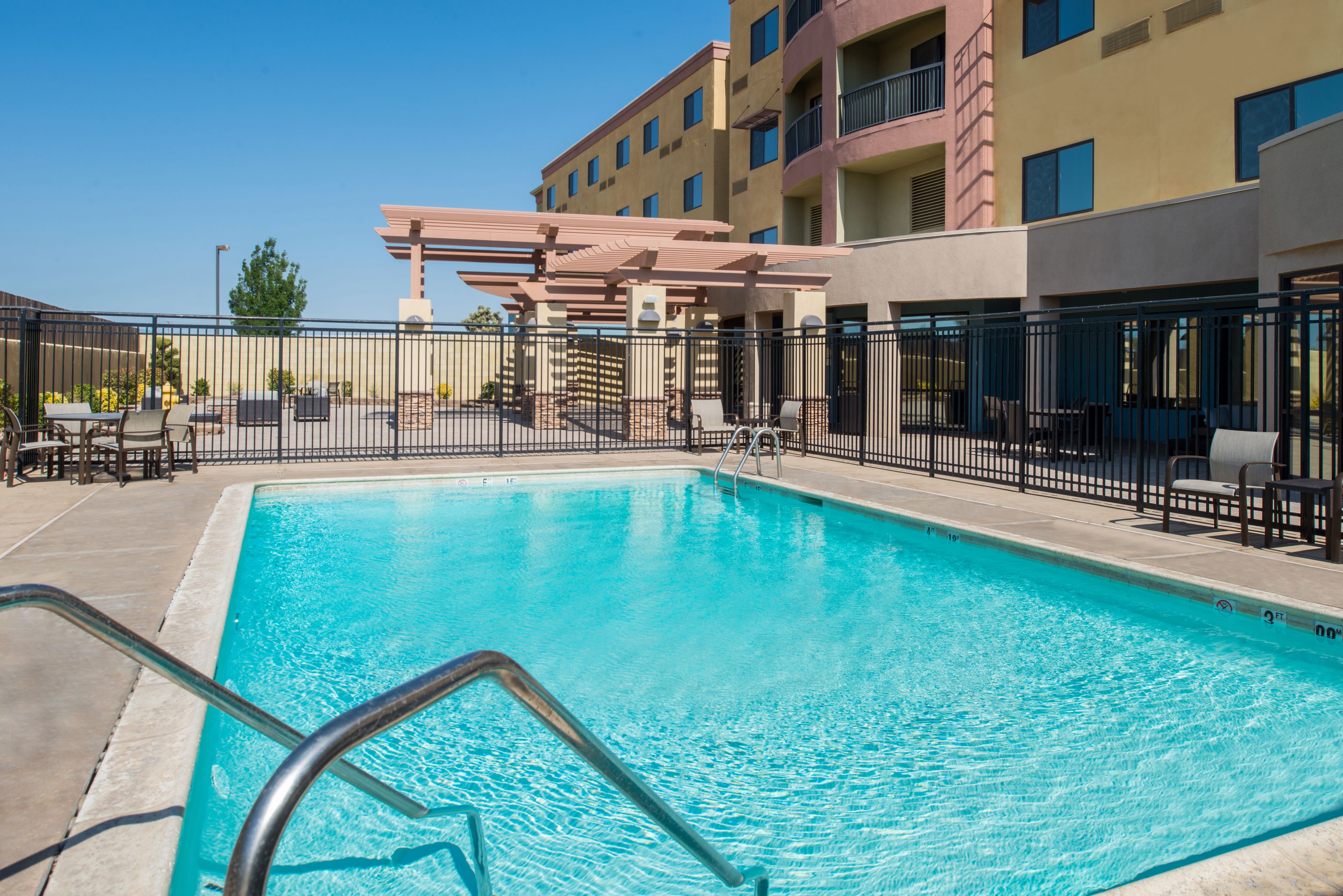 A swimming pool with a metal ladder, surrounded by a fence, and adjacent to a building with balconies and windows.