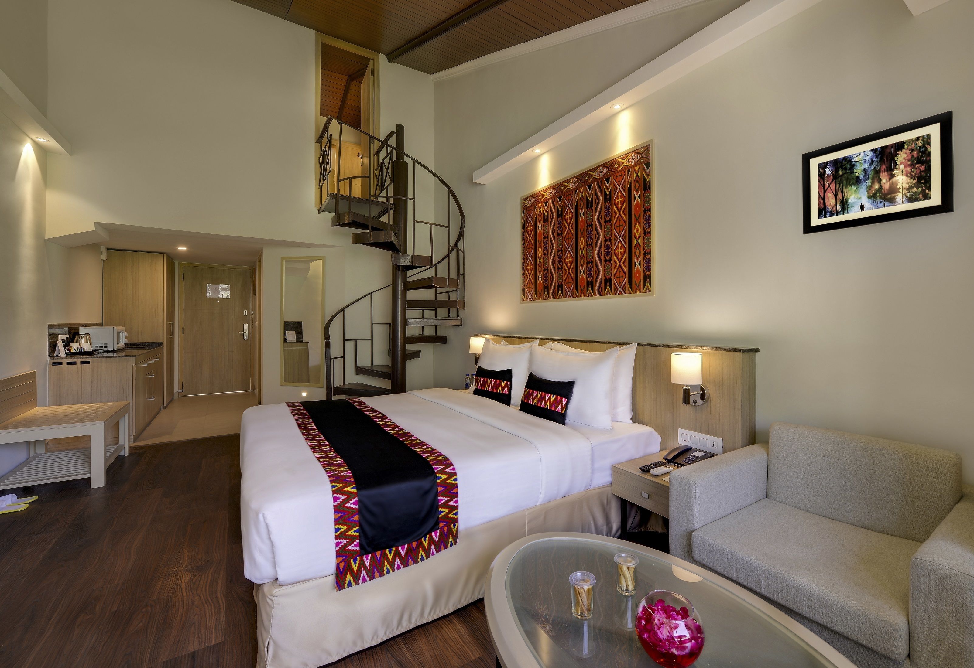 Habitación de hotel con cama, sofá, mesa de centro, escritorio, silla, escalera en espiral y una obra de arte en la pared.