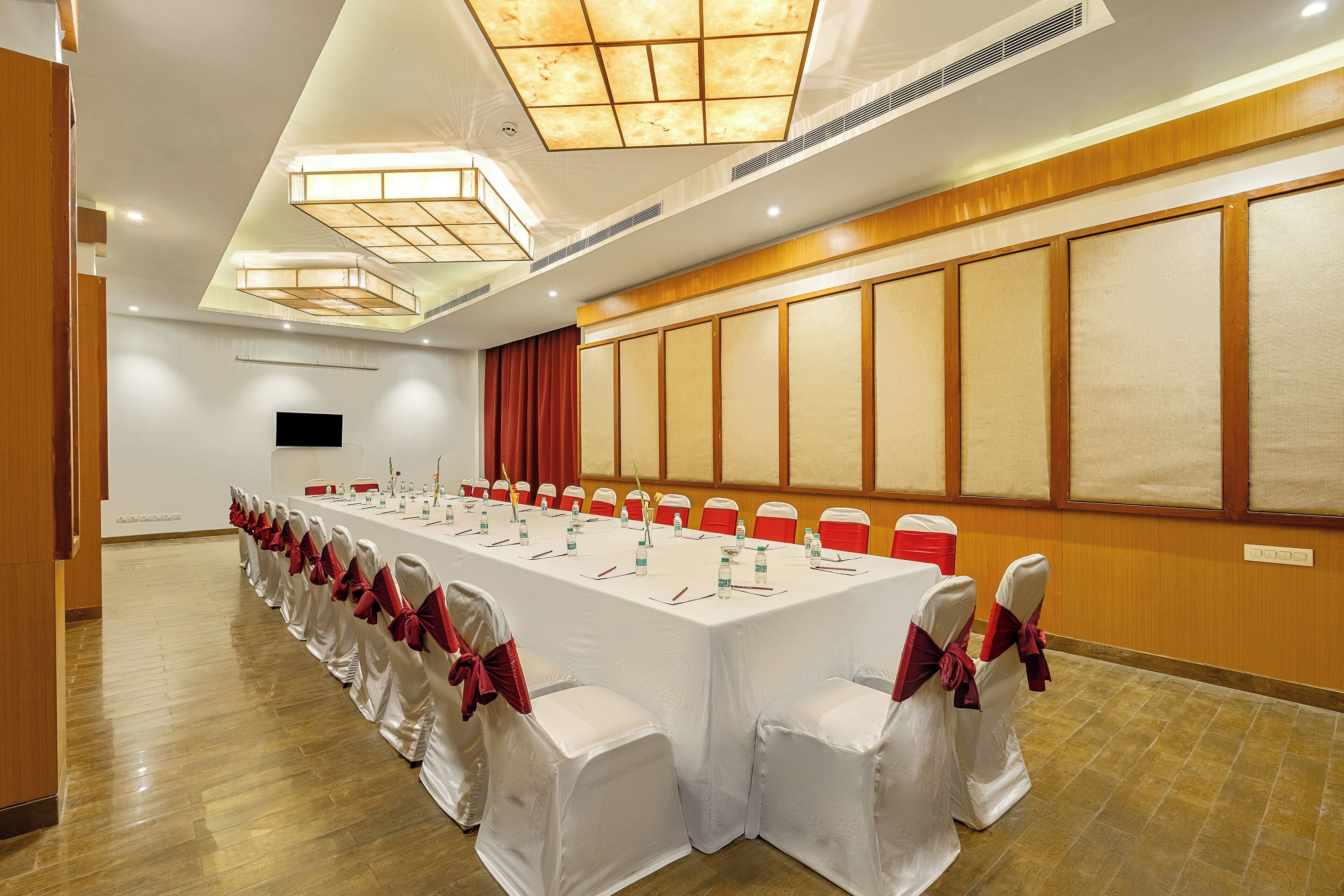 Un salón de conferencias con una larga mesa, sillas blancas con cintas rojas y un gran panel de pared de madera.