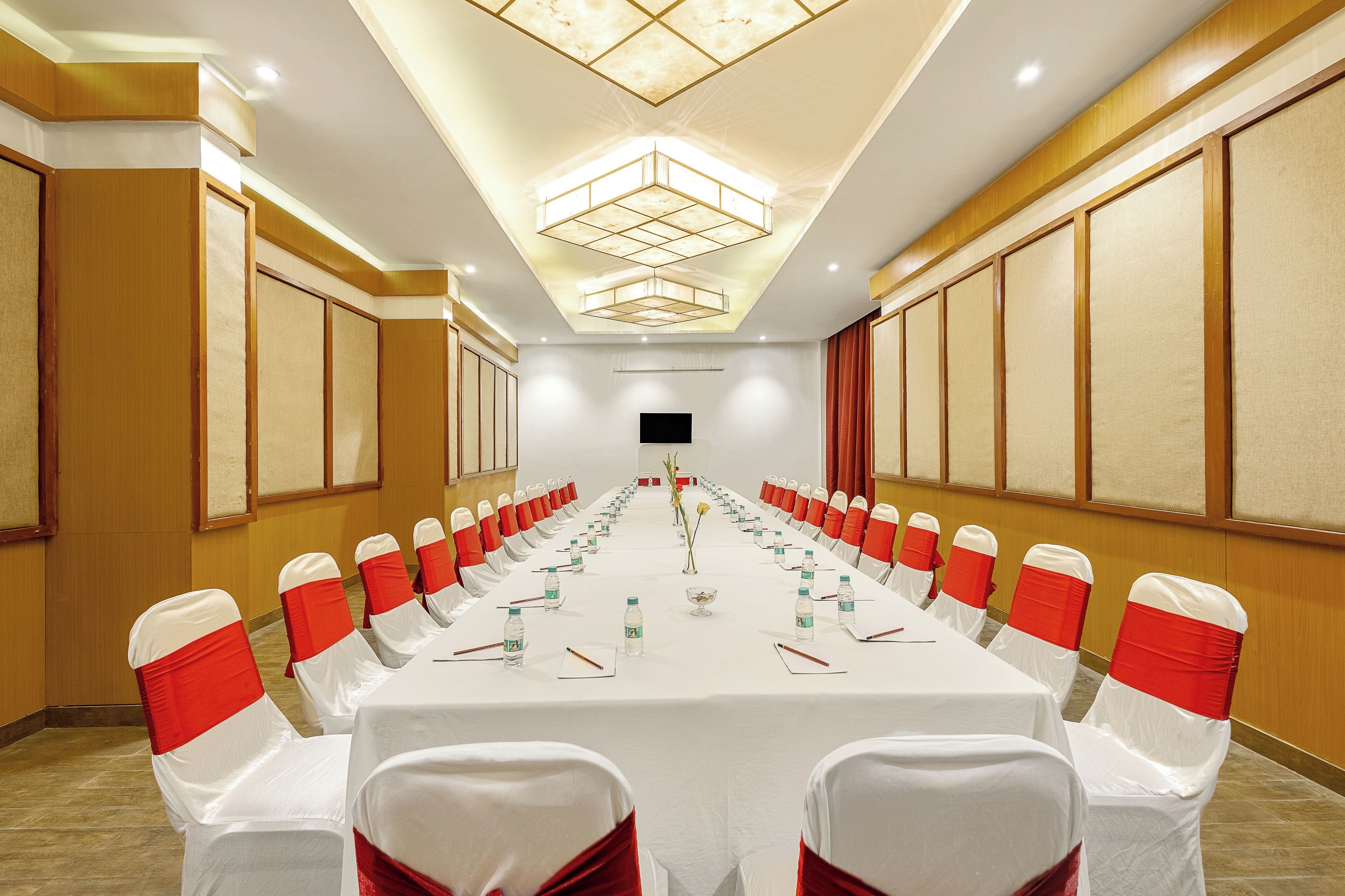 Une salle de conférence dotée d’une longue table, de chaises blanches ornées de rubans rouges et d’un plafond au design moderne.