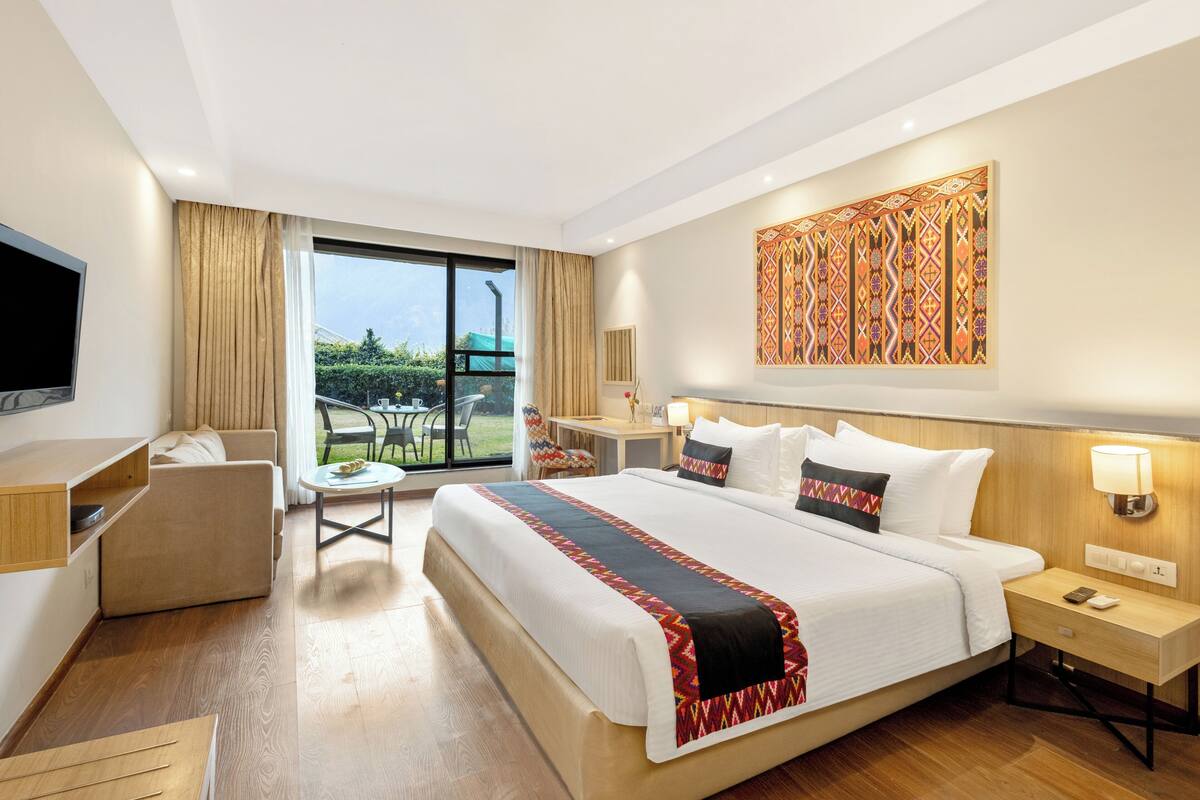 Ein Hotelzimmer mit einem großen Bett, einem Schreibtisch, einem Stuhl, einem Fernseher und einem Balkon mit Blick auf Grünflächen.